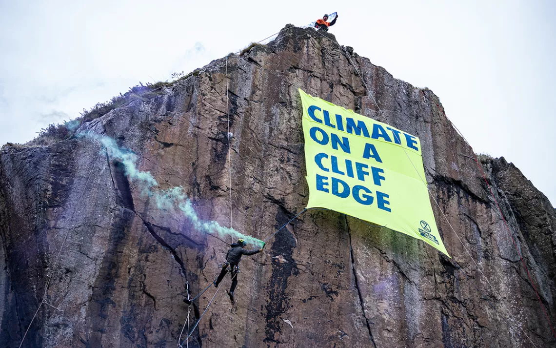 Experienced climbers scale a rock face near the historic Dumbarton castle in Glasgow, releasing a banner that reads “Climate on a Cliff Edge.” One activist, dressed as a globe, symbolically looms near the edge, while another plays the bagpipes on the shores below. | Photo courtesy of Extinction Rebellion and Mark Richards